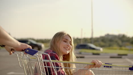 Young-romantic-couple-is-having-fun-with-supermarket-cart-in-the-evening.-Handsome-bearded-man-and-attractive-young-woman-are-spending-time-together-outdoors.