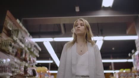 Pretty-lady-in-casual-clothes-is-walking-in-grocery-store-steering-shopping-trolley-with-food-inside-it-and-looking-around-at-shelves-with-products.-Women-and-shops-concept.
