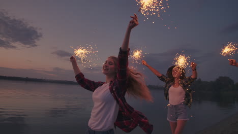 A-group-of-company-of-friends-men-and-women-Jolly-run-celebrate-the-holiday-on-the-beach-with-sparklers-and-fireworks.-Slow-motion-running-night-celebration-party-on-the-beach.