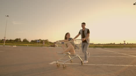 Young-stylish-coupe-having-fun-riding-with-shopping-cart-on-the-outdoor-parking-near-the-supermarket