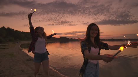 Cheerful-male-and-female-friends-are-running-along-the-beach-at-sunset-holding-sparkling-fireworks-and-runaway-lights-in-slow-motion.-Dancing-and-sunset-party-on-the-beach