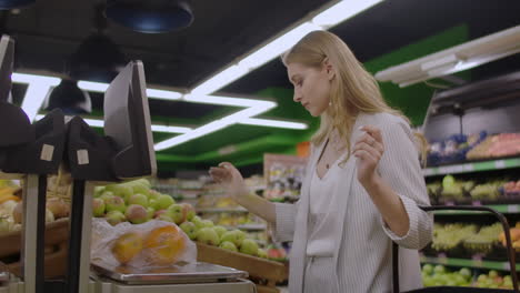 A-blonde-girl-in-a-supermarket-weighs-oranges-on-an-electronic-scale-pressing-the-display-standing-with-a-basket-in-her-hands