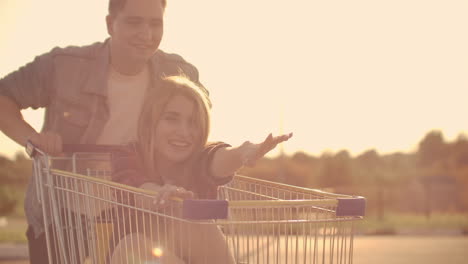 Side-view-of-a-young-man-and-woman-having-fun-outdoors-on-shopping-trolleys.-Multiethnic-young-people-racing-on-shopping-carts.-On-the-parking-zone-with-their