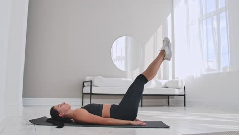 Sportswoman-training-at-home.-Fit-female-athlete-doing-toe-touch-single-arm-exercise-lying-on-floor-in-white-apartment.
