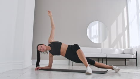 Young-sexy-brunette-woman-in-sportswear-doing-side-plank-exercise-with-a-tilt-at-home-in-a-white-interior.-Slow-motion