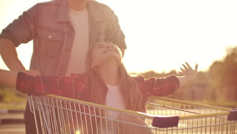 Couple-riding-with-shopping-cart-on-the-parking-outdoors.-Young-stylish-coupe-having-fun-riding-with-shopping-cart-on-the-outdoor-parking-near-the-supermarket