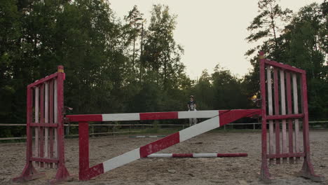 SLOW-MOTION-CLOSE-UP:-Young-horsegirl-horseback-riding-strong-brown-horse-jumping-the-fence-in-sunny-outdoors-sandy-parkour-riding-arena.-Competitive-rider-training-jumping-over-obstacles-at-sunset