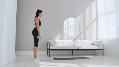 A-young-athletic-woman-in-a-bright-apartment-performs-a-deadlift-with-dumbbells-at-home-making-leans-forward-to-strengthen-the-muscles-of-the-thighs