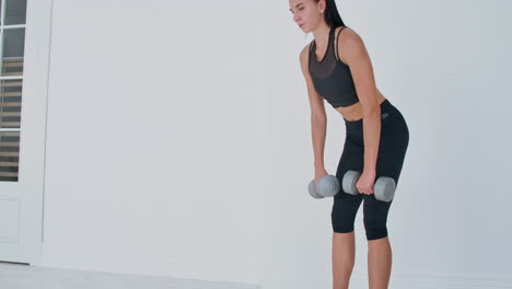A-young-athletic-woman-in-a-bright-apartment-performs-a-deadlift-with-dumbbells-at-home-making-leans-forward-to-strengthen-the-muscles-of-the-thighs