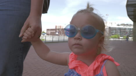 A-fair-haired-baby-girl-in-a-very-stylish-blue-round-sunglasses-walking-holding-mothers-hand
