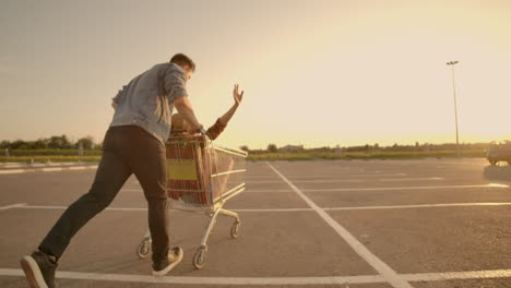 Back-view.-Cheerful-young-couple-in-love-man-and-woman-laughing-and-having-fun-while-riding-carts-in-supermarket-parking-in-slow-motion-at-sunset.