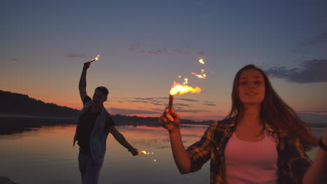 Young-happy-multiethnic-couple-holding-burning-sparkling-candles-and-running-by-the-sea-during-sunset.-Slow-motion-shot