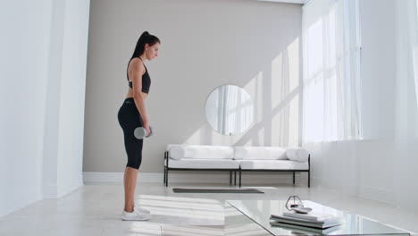 The-beautiful-female-performs-leans-forward-with-dumbbells-in-her-hands-in-the-white-interior-of-the-living-room.-In-slow-motion-to-do-exercises-on-the-muscles-of-the-thighs