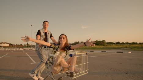 Young-romantic-couple-is-having-fun-with-supermarket-cart-in-the-evening.-Handsome-bearded-man-and-attractive-young-woman-are-spending-time-together-outdoors.