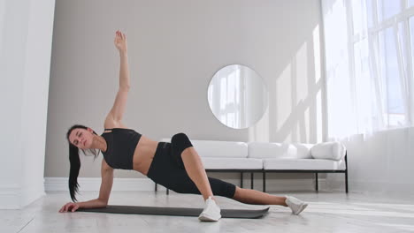 Young-sexy-brunette-woman-in-sportswear-doing-side-plank-exercise-with-a-tilt-at-home-in-a-white-interior.-Slow-motion