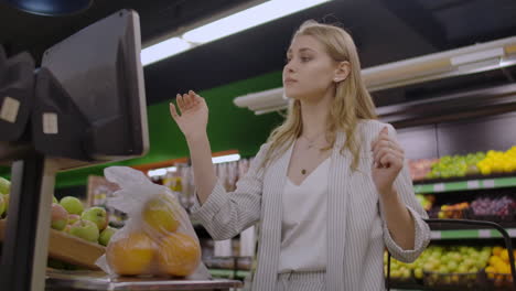 A-blonde-girl-in-a-supermarket-weighs-oranges-on-an-electronic-scale-pressing-the-display-standing-with-a-basket-in-her-hands
