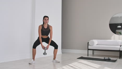 enduring-strong-determined-beautiful-concentrated-tanned-brunette-wearing-gray-stylish-sportive-top-and-leggings-woman-doing-deep-squats-with-dumbbells-in-outstretched-arms