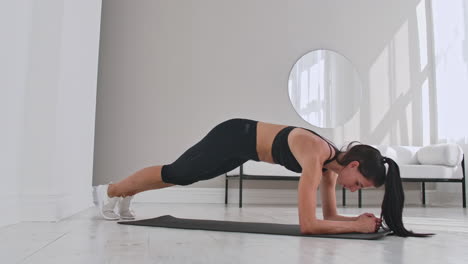 Female-brunette-fitness-trainer-demonstrating-plank-knee-to-chest-technique-on-a-mat-in-home