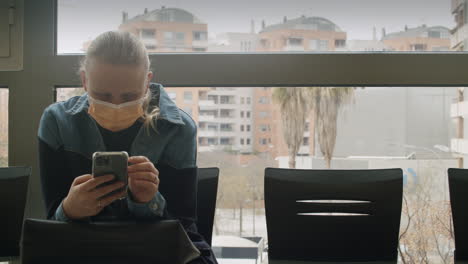 Woman-in-mask-using-smartphone-in-waiting-room