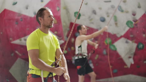 Portrait-of-beautiful-man-rock-climber-belaying-another-climber-with-rope.-Indoors-artificial-climbing-wall-and-equipment.
