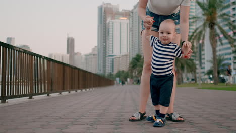 Happy-cute-baby-learns-to-walk-and-takes-first-steps-in-the-city-under-the-control-of-his-mom-holding-baby's-hands-in-the-background-of-the-city