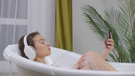 A-beautiful-brunette-woman-listens-to-music-in-headphones-on-a-smartphone-lies-in-a-bath-with-foam-and-rests-from-a-working-day.