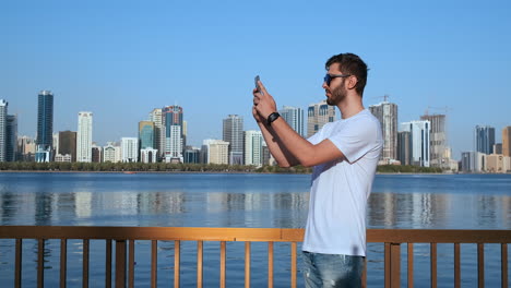 Happy-handsome-man-in-sunglasses-and-white-t-shirt-with-a-beard-taking-photos-on-a-smartphone-while-standing-on-the-waterfront-in-the-summer-town-in-the-background-and-buildings