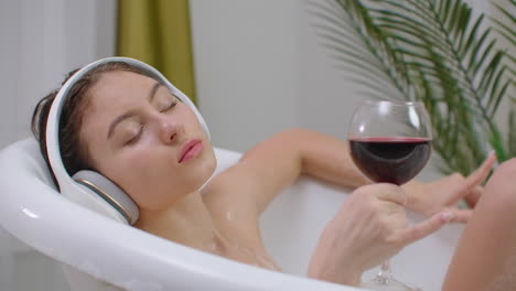 Relaxed-young-woman-listening-music-in-a-bubble-ba.-Close-up-of-a-relaxed-young-woman-listening-music-with-headphones-in-a-bubble-bath-and-drink-red-wine-from-a-glass