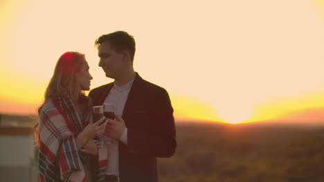 Young-married-couple-on-the-roof-hugging-and-drinking-red-wine-from-glasses-standing-dressed-in-plaid-and-admiring-the-beautiful-sunset-over-the-city..
