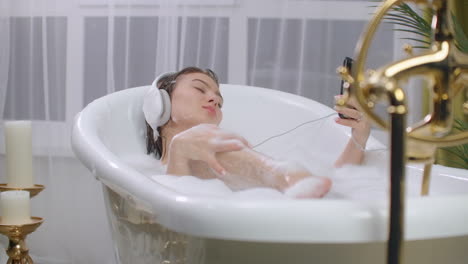 A-young-woman-lies-in-the-bath-wearing-headphones-and-looking-at-the-screen-of-a-smartphone.