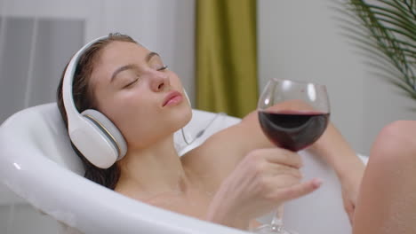 Beautiful-young-woman-enjoying-pleasant-bath-with-foam-holding-a-glass-of-wine-Woman-listening-to-music-in-bath.-Woman-listening-to-music-in-bubble-bath