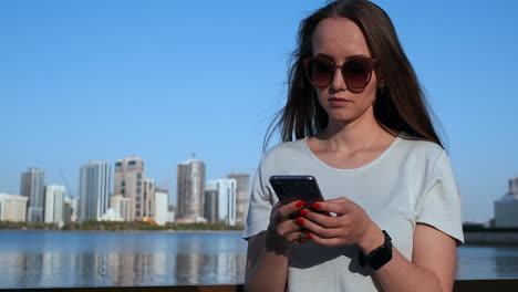 Schönes-Mädchen-Mit-Langen-Haaren-Und-Sonnenbrille-Nutzt-Smartphone-App-Am-Flusskai-Bei-Sonnenuntergang-In-Der-Nähe.