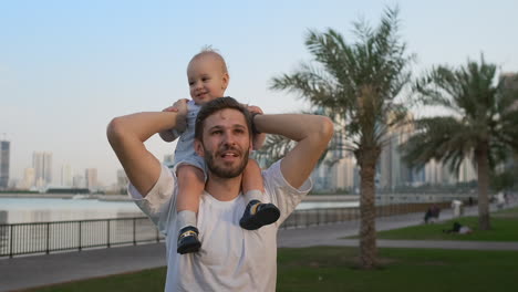 Loving-Father-in-a-white-t-shirt-in-the-summer-walks-with-a-child-sitting-on-the-neck-against-the-city-and-palm-trees