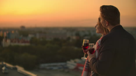 Man-and-young-pretty-woman-sitting-on-couch-holding-wine-glasses-and-kissing-on-rooftop-terrace-at-sunset
