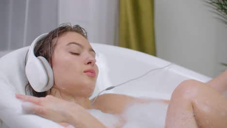 Young-woman-Relax-and-have-fun-dancing-hand-lying-in-a-bubble-bath-and-listening-to-music-through-headphones.