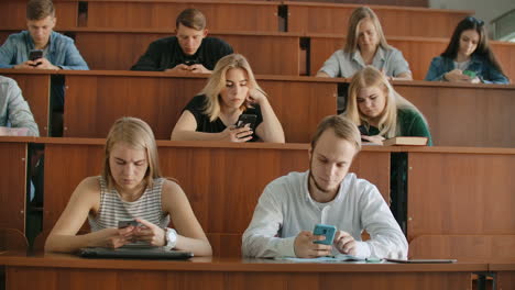 A-group-of-men-and-women-in-a-large-university-audience-silently-look-at-the-screens-of-smartphones-and-write-chat-messages.-Each-person-looks-at-the-screens-of-his-device-without-paying-attention-to-the-events-of-the-real-world.