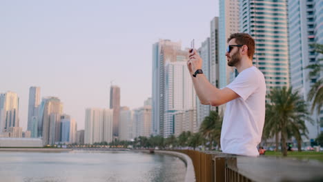 A-young-man-in-a-white-t-shirt-standing-on-the-waterfront-against-the-background-of-the-modern-city-takes-photos-and-videos-on-the-phone