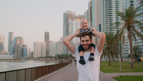 A-young-father-in-a-white-t-shirt-and-bristles-hipster-walking-with-his-son-sitting-on-his-neck-on-the-waterfront-against-the-background-of-the-modern-city-in-the-summer-on-the-waterfront