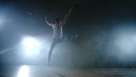 A-modern-ballet-a-man-performs-jumps-and-spins-in-the-light-of-spotlights-and-smoke-on-a-dark-background.-Acrobatic-choreography-rehearsal-of-the-script-of-modern-ballet.