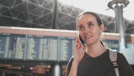 Young-beautiful-woman-standing-at-the-airport-calls-on-the-phone-on-the-background-of-the-scoreboard-with-information-about-the-departures.