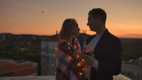 Young-married-couple-on-the-roof-hugging-and-drinking-red-wine-from-glasses-standing-dressed-in-plaid-and-admiring-the-beautiful-sunset-over-the-city..