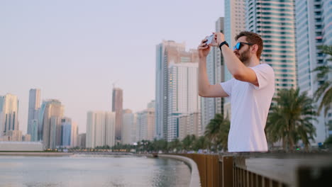 A-young-man-in-a-white-t-shirt-standing-on-the-waterfront-against-the-background-of-the-modern-city-takes-photos-and-videos-on-the-phone-for-social-networks-is-live