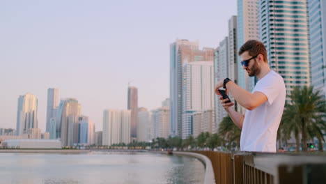 Happy-handsome-man-in-sunglasses-and-white-t-shirt-with-a-beard-taking-photos-on-a-smartphone-while-standing-on-the-waterfront-in-the-summer-town-in-the-background-and-buildings