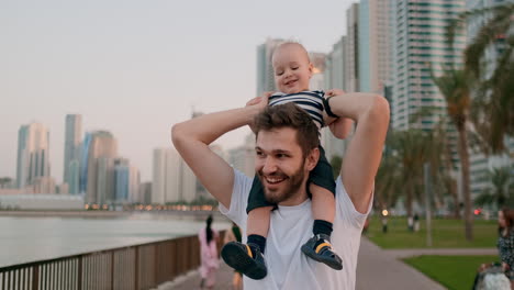 Happy-young-family-loving-Young-father-in-white-t-shirt-and-bristles-hipster-walking-with-his-son-sitting-on-his-neck-on-the-waterfront-in-the-background-of-modern-city-in-summer-on-the-waterfront