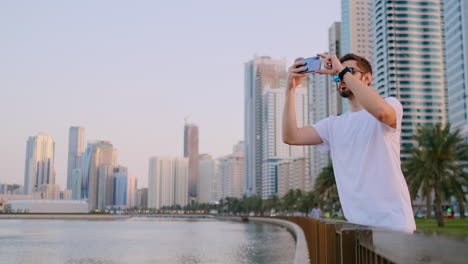 A-young-male-tourist-in-sunglasses-and-bristles-hipster-in-a-white-t-shirt-standing-on-the-waterfront-against-the-background-of-the-modern-city-takes-photos-and-videos-on-the-phone-for-social-networks-is-live-broadcast