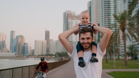 Smiling-Young-father-in-white-t-shirt-and-bristles-hipster-walking-with-his-son-sitting-on-the-neck-on-the-waterfront-in-the-background-of-the-modern-city-in-the-summer-on-the-waterfront