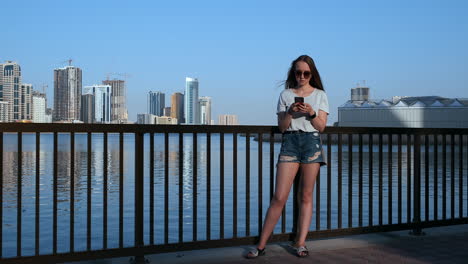 Beautiful-girl-with-long-hair-in-sunglasses-using-smartphone-app-at-sunset-river-quay-near.