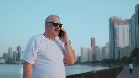Happy-handsome-senior-man-in-sunglasses-and-white-t-shirt-talking-on-the-phone-standing-on-the-waterfront-in-summer-against-the-city-and-buildings