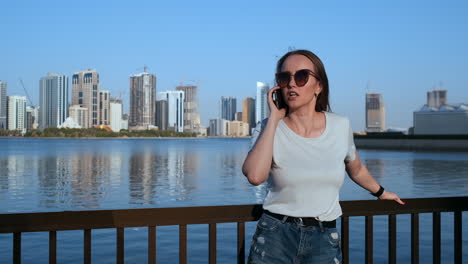 Happy-beautiful-woman-in-sunglasses-and-white-t-shirt-talking-on-the-phone-standing-on-the-promenade-in-the-summer-against-the-city-and-buildings