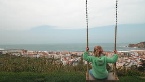 Kleines-Mädchen-Blickt-Vom-Swing-Nazare-In-Portugal-Auf-Das-Meer-Und-Den-Ferienort
